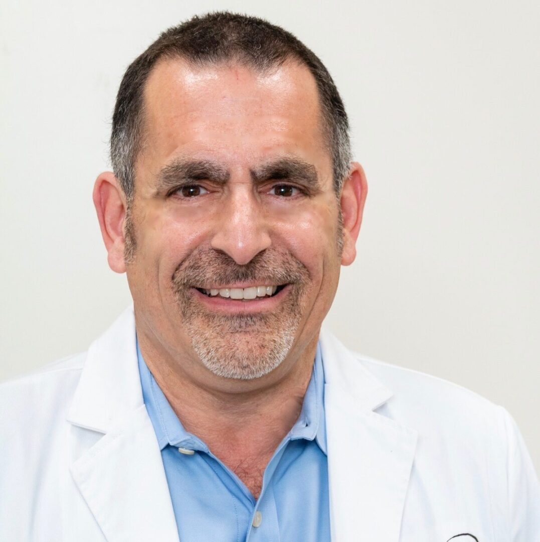 A man in white coat and blue shirt smiling.